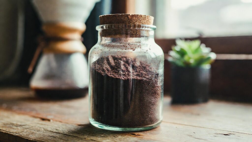 ガラス容器に保存された乾燥コーヒーかすと、背景に置かれたコーヒードリッパー